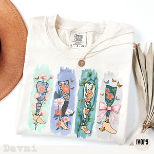 May include: Ivory-colored t-shirt featuring a floral prosthetic leg design. The design includes four vertical panels with different floral and butterfly arrangements. The shirt has a Comfort Colors tag and a gold necklace. The word "Ivory" is printed at the bottom right.