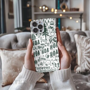 May include: A white phone case with a green illustrated design featuring trees, a car, and the word "HOA" repeated. The word "Forever" is printed at the bottom of the case.