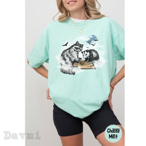 K&ouml;nnte beinhalten: Ein t&uuml;rkisfarbenes T-Shirt mit einem Aufdruck eines Waschb&auml;ren und eines Opossums, die auf einer Wolke sitzen, mit einem Ouija-Brett vor ihnen. Der Aufdruck ist in Schwarzwei&szlig;.