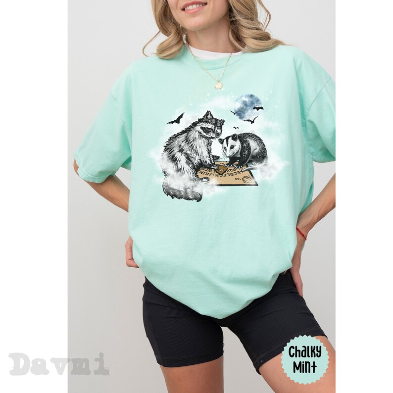 K&ouml;nnte beinhalten: Ein t&uuml;rkisfarbenes T-Shirt mit einem Aufdruck eines Waschb&auml;ren und eines Opossums, die auf einer Wolke sitzen, mit einem Ouija-Brett vor ihnen. Der Aufdruck ist in Schwarzwei&szlig;.