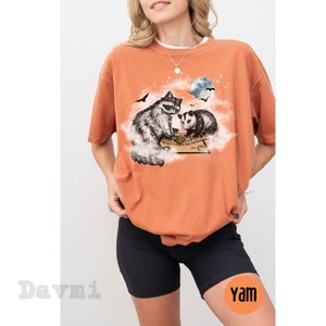 K&ouml;nnte beinhalten: Eine Frau tr&auml;gt ein orangefarbenes T-Shirt mit einem Aufdruck eines Waschb&auml;ren und eines Opossums, die auf einer Wolke mit einem Ouija-Brett darunter sitzen. Das Shirt ist &uuml;berdimensioniert und hat kurze &Auml;rmel. Die Frau tr&auml;gt auch schwarze Radlerhosen.