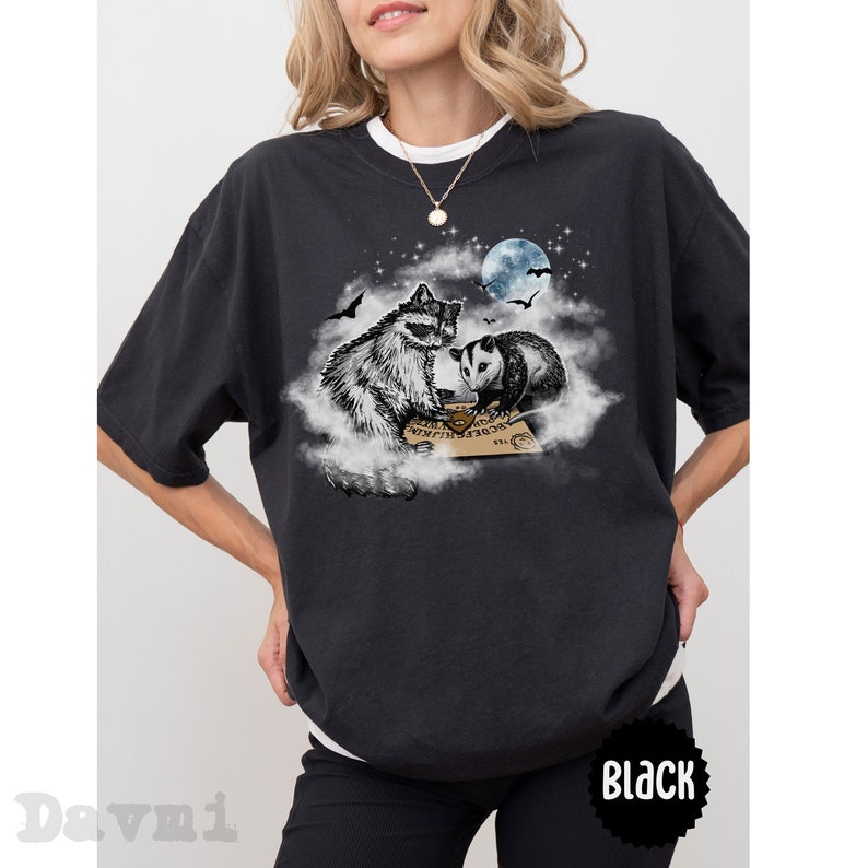K&ouml;nnte beinhalten: Ein schwarzes T-Shirt mit einem Aufdruck eines Waschb&auml;ren und eines Opossums, die auf einer Wolke sitzen, mit einem Ouija-Brett vor ihnen. Der Aufdruck ist vor dem Hintergrund eines sternenklaren Nachthimmels mit einer Sichelmond.