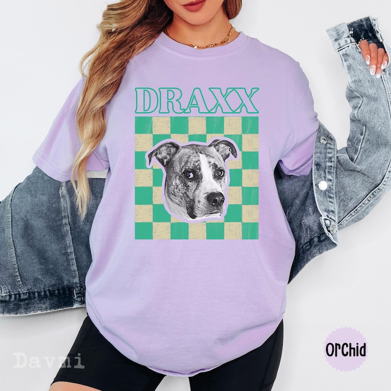 Peut inclure: Un t-shirt violet clair avec le texte DRAXX en sarcelle au-dessus d'un carr&eacute; &agrave; damier vert et beige. Au centre du carr&eacute; se trouve un portrait photographique en noir et blanc d'un chien avec une tache blanche sur le museau et des oreilles sombres.