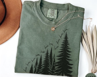 Peut inclure: Un t-shirt à col rond vert olive avec un motif noir représentant des pins et le texte cursif "hoa" répété en diagonale ascendante. Le t-shirt porte une étiquette de marque Comfort Colors à l'intérieur du col et un collier doré.