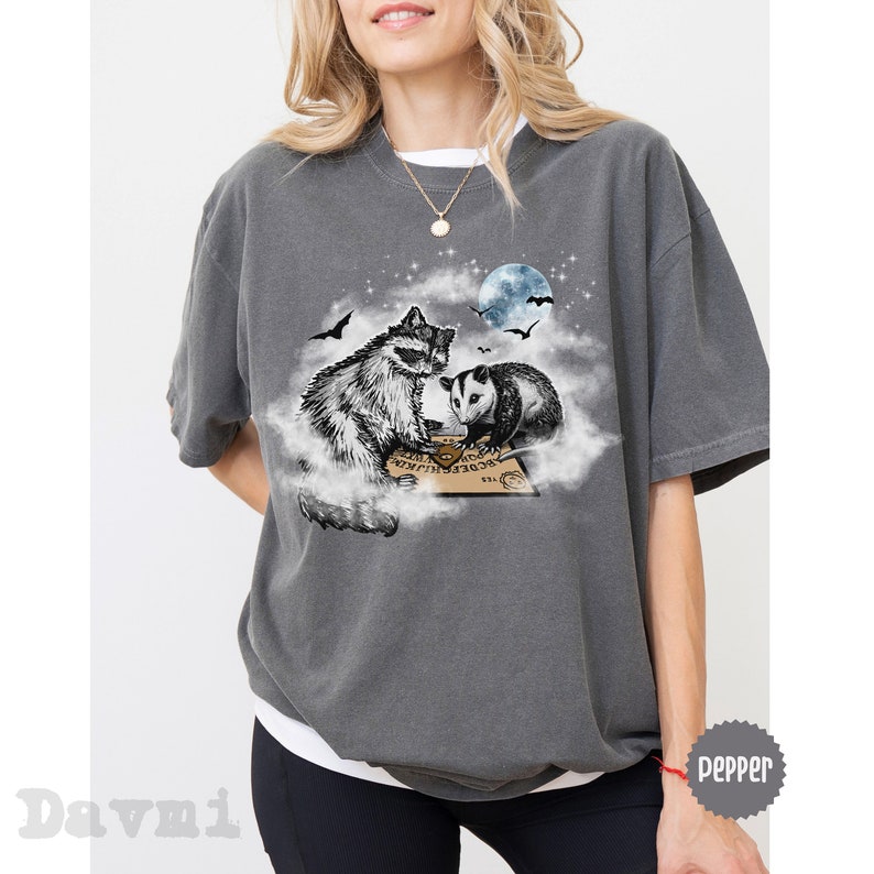 K&ouml;nnte beinhalten: Ein graues T-Shirt mit einem Aufdruck eines Waschb&auml;ren und eines Opossums, die auf einer Wolke sitzen, mit einem Ouija-Brett vor ihnen. Der Aufdruck ist in Schwarzwei&szlig; mit einem blauen Mond und Sternen im Hintergrund.