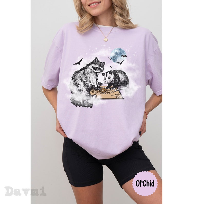 K&ouml;nnte beinhalten: Ein lavendelfarbenes T-Shirt mit einem grafischen Aufdruck eines Waschb&auml;ren und eines Opossums, die an einem Ouija-Brett sitzen, mit einem Vollmond im Hintergrund.