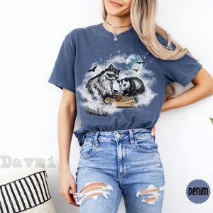 K&ouml;nnte beinhalten: Ein marineblaues T-Shirt mit einem Aufdruck eines Waschb&auml;ren und eines Opossums, die auf einer Wolke sitzen, mit einem Ouija-Brett vor ihnen. Der Mond und die Sterne sind im Hintergrund.