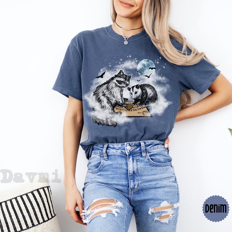 K&ouml;nnte beinhalten: Ein marineblaues T-Shirt mit einem Aufdruck eines Waschb&auml;ren und eines Opossums, die auf einer Wolke sitzen, mit einem Ouija-Brett vor ihnen. Der Mond und die Sterne sind im Hintergrund.