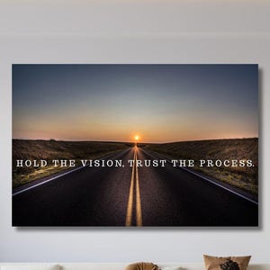 Op de afbeelding: Een rechthoekige wandkunstprint met een landschapsfoto van een lange, rechte asfaltweg die naar een zonsondergang aan de horizon leidt. De tekst HOLD THE VISION, TRUST THE PROCESS staat in witte hoofdletters in het midden gedrukt.