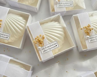 Puede incluir: Jabones blancos en forma de concha en cajas transparentes, atados con cintas blancas. Cada caja está decorada con flores secas y una perla. El texto en la cinta está en turco.