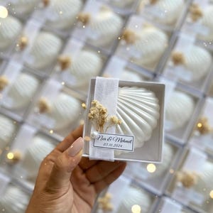 May include: White seashell-shaped soap favours in clear boxes, ideal for a wedding. Each soap is decorated with a small floral accent and a label reading "Nur & Mehmet 03.10.2024".
