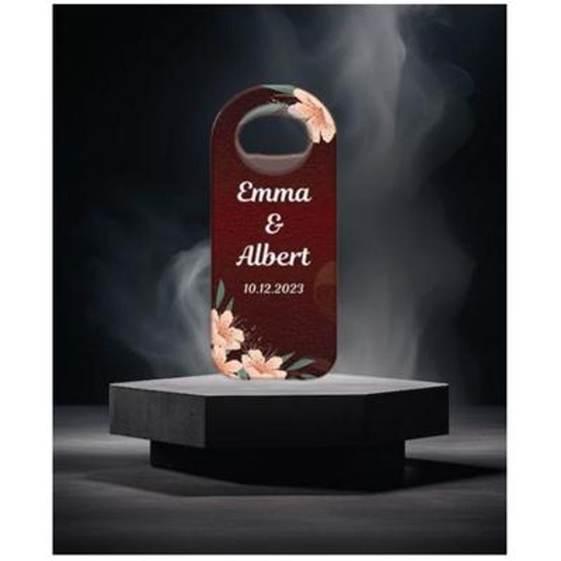 May include: A maroon door hanger with a floral design and the names "Emma & Albert" along with the date "10.12.2023" in white text. The door hanger is displayed on a black pedestal against a dark background.