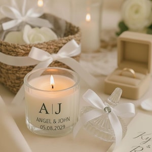 May include: A lit candle in a clear glass holder with the initials "A|J", the names "ANGEL & JOHN", and the date "05.08.2029". A small ring box and a decorative glass jar with a white bow are also visible. A woven basket with white flowers and candles is in the background.