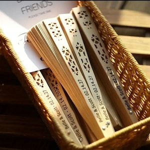 May include: A collection of light brown wooden fans with intricate cut-out designs, arranged in a woven basket. Each fan is engraved with "Belle & Scott 02.14.27" and "Belle & Scott 02.14.2022". A card with the word "FRIENDS" is visible.