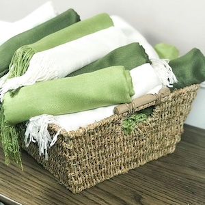 May include: A wicker basket containing folded green and white scarves. The scarves are in various shades of green, with fringed edges. A wooden handle is attached to the basket.