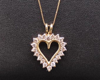Puede incluir: Un collar con colgante en forma de corazón de oro que presenta un diseño central de corazón abierto rodeado por un halo de pequeñas piedras claras de corte redondo. El colgante cuelga de una delicada cadena de oro de eslabones cuadrados.