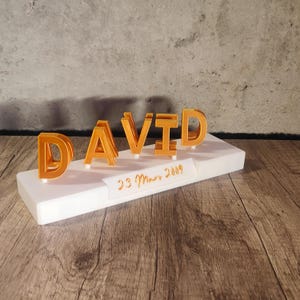 Peut inclure: Un socle rectangulaire blanc avec le nom "DAVID" écrit en lettres dorées. La date "23 Mars 2009" est inscrite sur le socle.