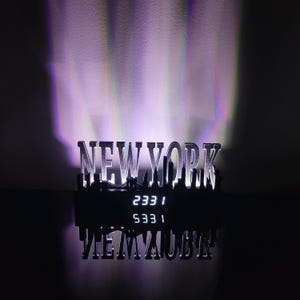 Peut inclure: Un panneau "NEW YORK" argenté avec une silhouette de paysage urbain, éclairé par une lumière violette. Le panneau affiche l'heure "2331" et "5331". Le reflet du panneau est visible en dessous.