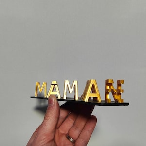 Peut inclure: Un signe "MAMAN" doré sur une base noire. Les lettres sont en 3D, et le signe est tenu par une main. Le fond est de couleur gris clair uni.