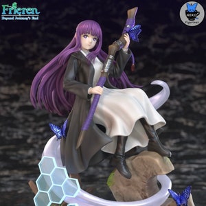 May include: A detailed figurine of a character from "Frieren: Beyond Journey's End." The figure features a woman with long purple hair, wearing a black coat over a white dress, and holding a staff adorned with butterflies. The base includes a hexagonal design and a rock-like structure.