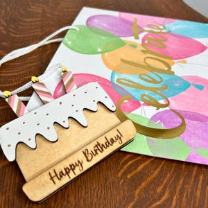 May include: A wooden cake with white frosting and sprinkles, with the words "Happy Birthday!" engraved on the bottom. The cake is attached to a white gift bag with a gold inscription that says "Celebrate" and colorful balloons.