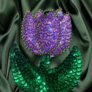 Puede incluir: Una aplicación de flor de lentejuelas moradas y verdes. La flor es un tulipán con un centro morado y hojas verdes. Las lentejuelas son iridiscentes y brillan.