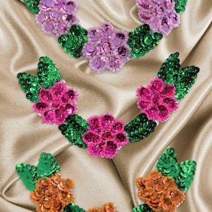 May include: Three floral appliques with green leaves and pink, fuchsia, and orange flower centers. The appliques are made of sequins and are arranged in a crescent shape.