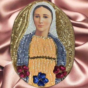 May include: Oval-shaped religious icon featuring a portrait of a woman with a halo. The icon is embellished with gold, blue, and red sequins. The background is a pink satin fabric.