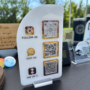 Może przedstawiać: Biały znak akrylowy z złotymi akcentami i czterema kodami QR. Znak zawiera tekst "FOLLOW US", "TEXT US", "PAY US =)" oraz ikony dla Instagrama, WhatsApp i znaku dolara.