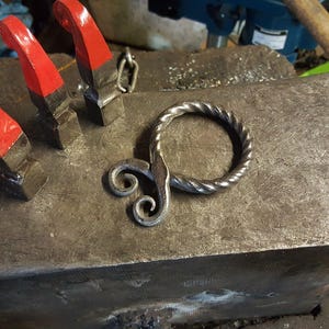 May include: A handcrafted metal ring with a twisted design and decorative swirls, resting on a weathered anvil. Three red-handled clamps are positioned nearby. The ring is likely a piece of blacksmithing art.