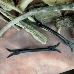 Puede incluir: Dos ganchos de metal negro con un diseño único y puntiagudo. Los ganchos están sobre una superficie marrón claro.