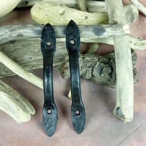 Puede incluir: Dos tiradores de cajón de metal negro, alargados, con un acabado texturizado y un agujero en cada extremo. Los tiradores están colocados verticalmente sobre un fondo de madera flotante y una superficie marrón rosada.