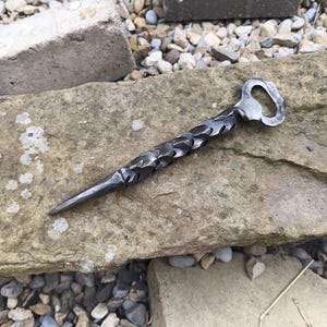 May include: A hand-forged bottle opener with a twisted metal design. The opener has a pointed end and a bottle opener at the other end.