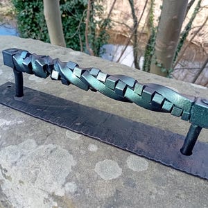 May include: A black metal door handle with a twisted, square-shaped design. The handle has a dark green patina finish.
