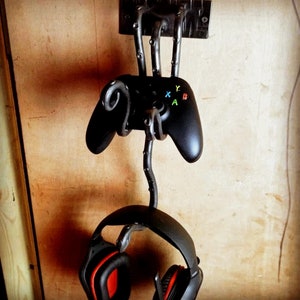 May include: A black metal wall mount with a curved design holds a black video game controller and a black headset. The controller has green, yellow, and red buttons labelled X, Y, A, and B.