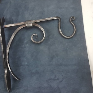 Puede incluir: Un soporte de pared de metal con un diseño decorativo de volutas. El soporte tiene un solo gancho en el lado derecho y una base plana en el lado izquierdo.