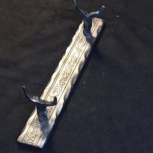 May include: A silver metal stand with two curved arms for holding swords or other items. The stand has an ornate design with a repeating pattern.