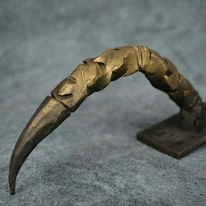 May include: A gold-colored metal door handle shaped like a claw. The handle is curved and has a sharp point at the end.