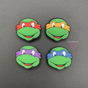 May include: Four green Teenage Mutant Ninja Turtle face charms with red, orange, purple, and blue masks. Each charm has a smiling mouth and white eyes. The text "CHARM UP" is visible in the upper right corner.