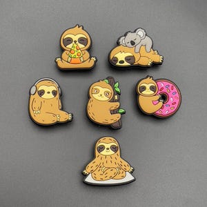 Könnte beinhalten: Ein Set aus sechs bunten Schuhanhängern aus Gummi mit Cartoon-Faultieren in verschiedenen Posen. Ein Faultier isst Pizza, eines schläft mit einem Koala, eines trägt Kopfhörer, eines klammert sich an einen Ast, eines umarmt einen rosa glasierten Donut und eines sitzt im Lotussitz.