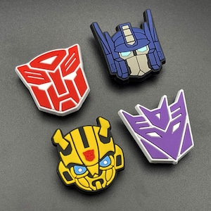 May include: Four shoe charms featuring Transformers characters and symbols. One is a red Autobot logo, one is a purple Decepticon logo, one is a blue and grey Optimus Prime head, and one is a yellow and black Bumblebee head with blue eyes.