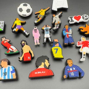 May include: A collection of colorful soccer-themed charms, including soccer balls, goal posts, and figurines of soccer players in various uniforms. The charms are in various colors, including white, red, blue, yellow, and green.