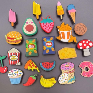 Può includere: Una collezione di ciondoli colorati a tema cibo, tra cui gelati, ghiaccioli, un hamburger, avocado, fetta di pizza e ciambelle. Altri ciondoli includono popcorn, patatine, un fungo e una banana. I ciondoli sono realizzati in materiale lucido.