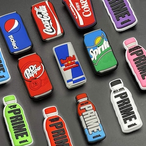 May include: A collection of drinks-themed charms featuring popular brands like Coca-Cola, Pepsi, and Sprite, alongside Red Bull and Dr. Pepper can designs. Also includes Prime drink bottle charms. The charms are displayed on a dark background.