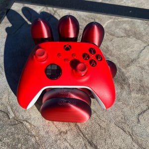 May include: A red Xbox controller rests on a 3D-printed hand-shaped stand. The controller features black buttons and white accents. The stand is also red and has a textured appearance, with the shadow of the hand visible.