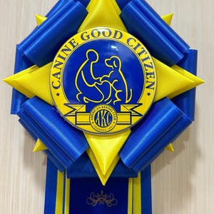 May include: A blue and yellow rosette ribbon with the text "CANINE GOOD CITIZEN" and "AKC" on a circular emblem. The ribbon has a blue and yellow starburst design and a long blue ribbon tail.