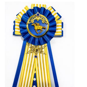 May include: A blue and gold equestrian award ribbon. The ribbon features a gold horse and rider emblem, blue and white striped ribbons, and gold filigree accents. The ribbon is approximately 8 inches in length.