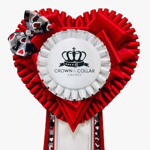May include: A heart-shaped red ribbon award with a white and black bow. The award features a white circular emblem with a crown logo and the text "CROWN & COLLAR AWARDS". The ribbon has a white section at the bottom.