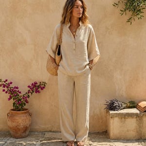 Op de afbeelding: Een beige linnen tweedelige set bestaande uit een losvallend overhemd met korte mouwen, een kraag en knoopsluiting, en een bijpassende broek met wijde pijpen. De outfit wordt gecombineerd met een gevlochten strooien schoudertas met een beige leren riem.