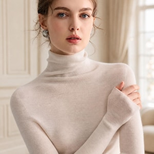 May include: A long-sleeved, cream-colored turtleneck sweater featuring a fitted silhouette and ribbed cuffs. The knit fabric has a smooth, fine texture, and the high collar extends to the base of the neck, providing a clean and simple appearance.
